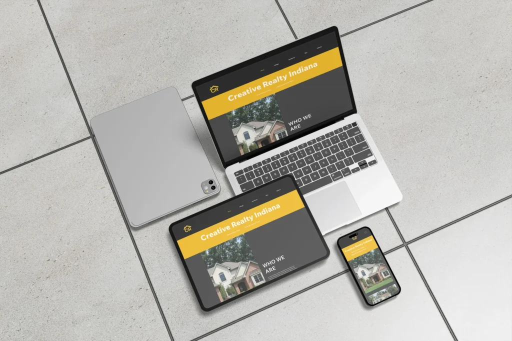 Responsive web design mockup of the Creative Realty Indiana website displayed on a laptop, tablet, and smartphone arranged on a tiled surface — dark charcoal and yellow color scheme with 'Creative Realty Indiana' heading and a house photo