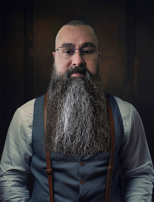 Eric Smoldt, graphic artist and visual communications designer, photographed against a dark wood-panel background wearing a slate vest, white shirt, and brown leather suspenders.