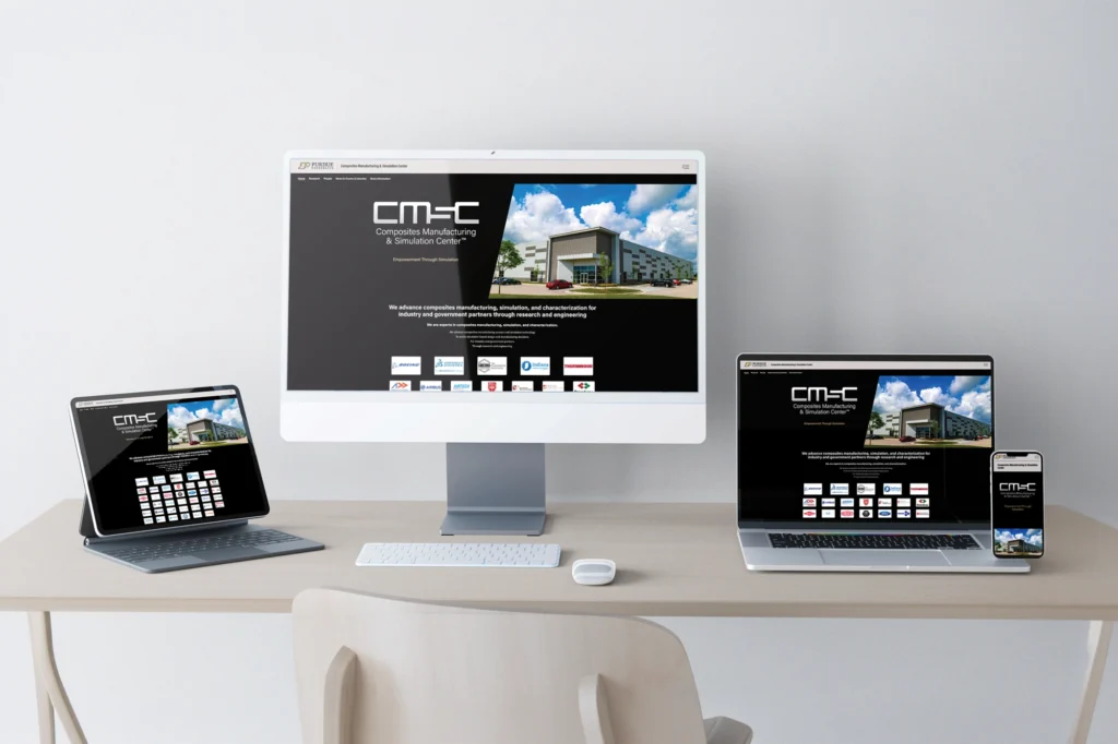 Responsive web design mockup for Purdue University's Composites Manufacturing and Simulation Center (CMSC), displayed on a desktop monitor, tablet, laptop, and phone on a light wood desk. The dark-themed homepage shows the CMSC logo, a photo of the center's building, the tagline about advancing composites manufacturing and simulation, and a grid of industry partner logos including Boeing and Airbus.